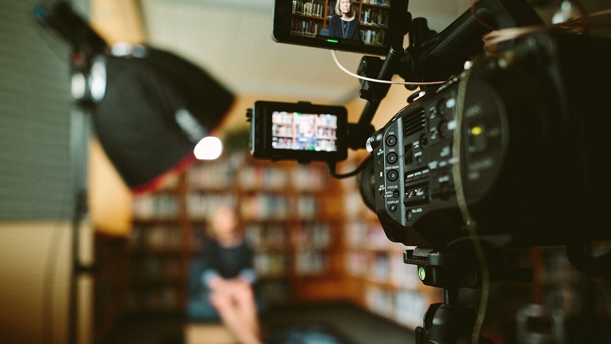 Kamera im Vordergrund filmt eine Interviewsituation vor einem Bücherregal