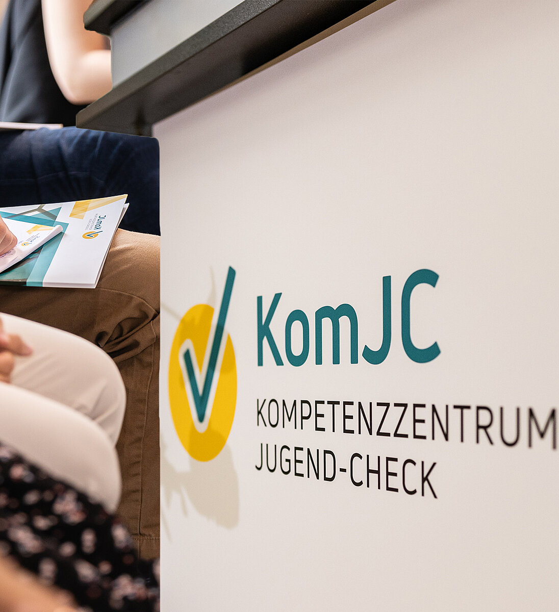 Logo des Kompetenzzentrums Jugend-Check neben sitzenden Teilnehmenden bei einer Beteiligungsveranstaltung 