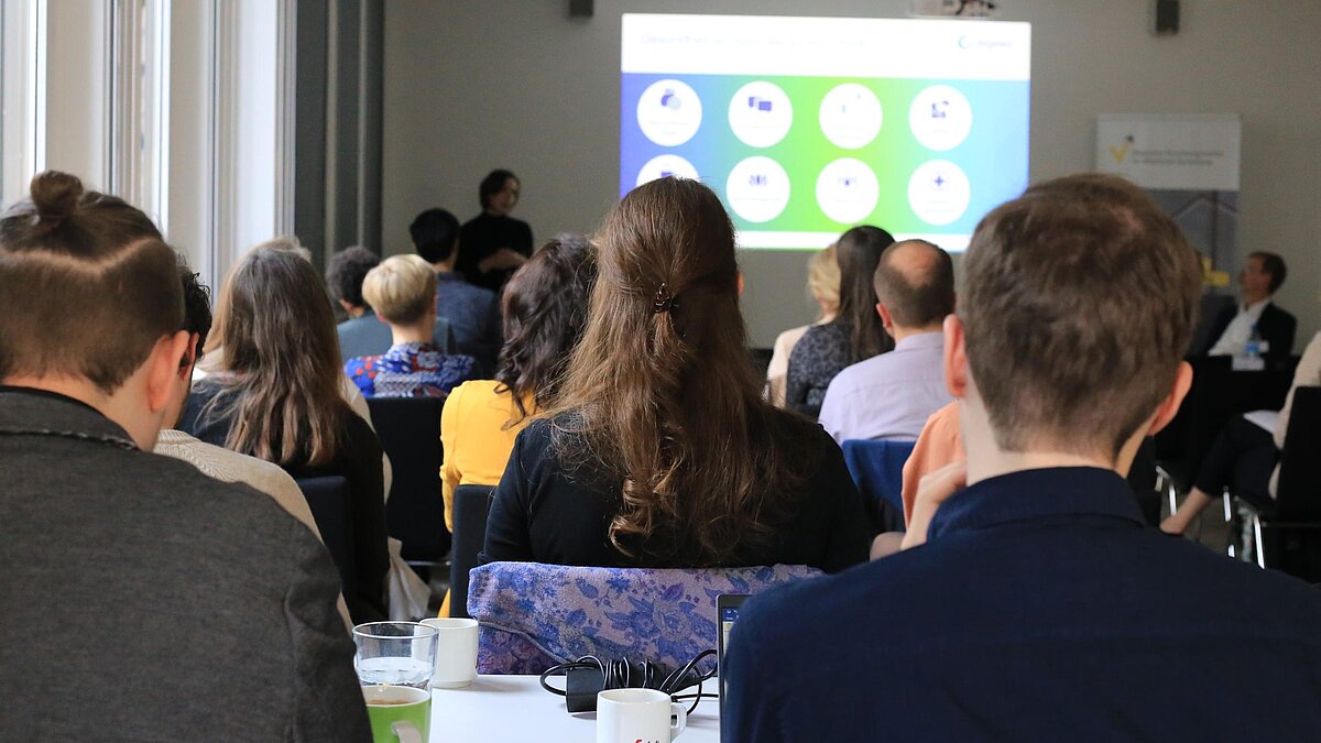 von hinten aufgenommene sitzende Workshopteilnehmende, im Vorderung ist eine referierende Person vor einem Beamerbild zu sehen