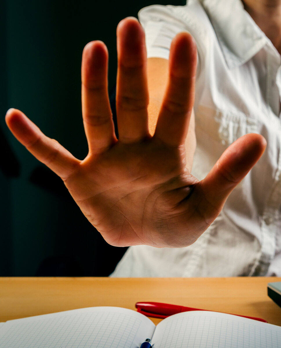 Eine Frau mit erhobener Hand und abwehrender Geste sitzt am Schreibtisch.