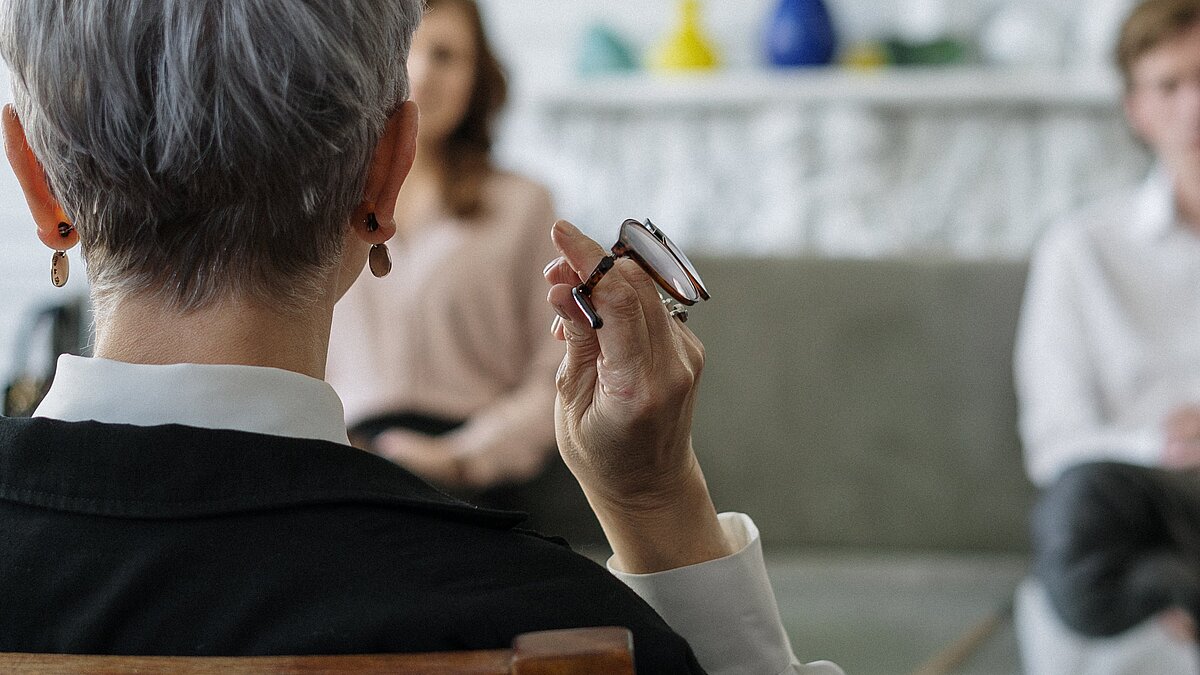 Eine Frau ist von hinten zu sehen. Sie hat eine Brille in der Hand und sitzt zwei Personen in einem Büro gegenüber.