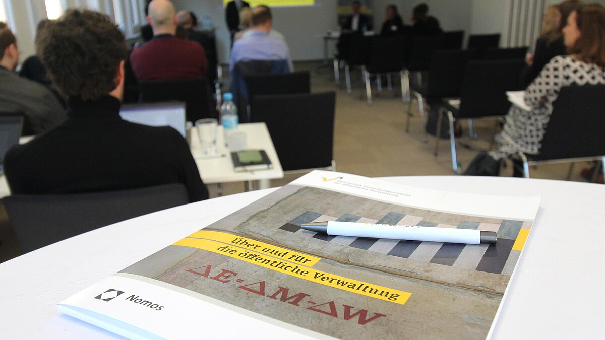 Im Vordegrund liegt eine FÖV-Tagungsmappe mit Kuli auf einem weißen Stehtisch. Im Hintergrund sitzen Tagungsteilnehmende auf schwarzen Stühlen und eine Person referiert.